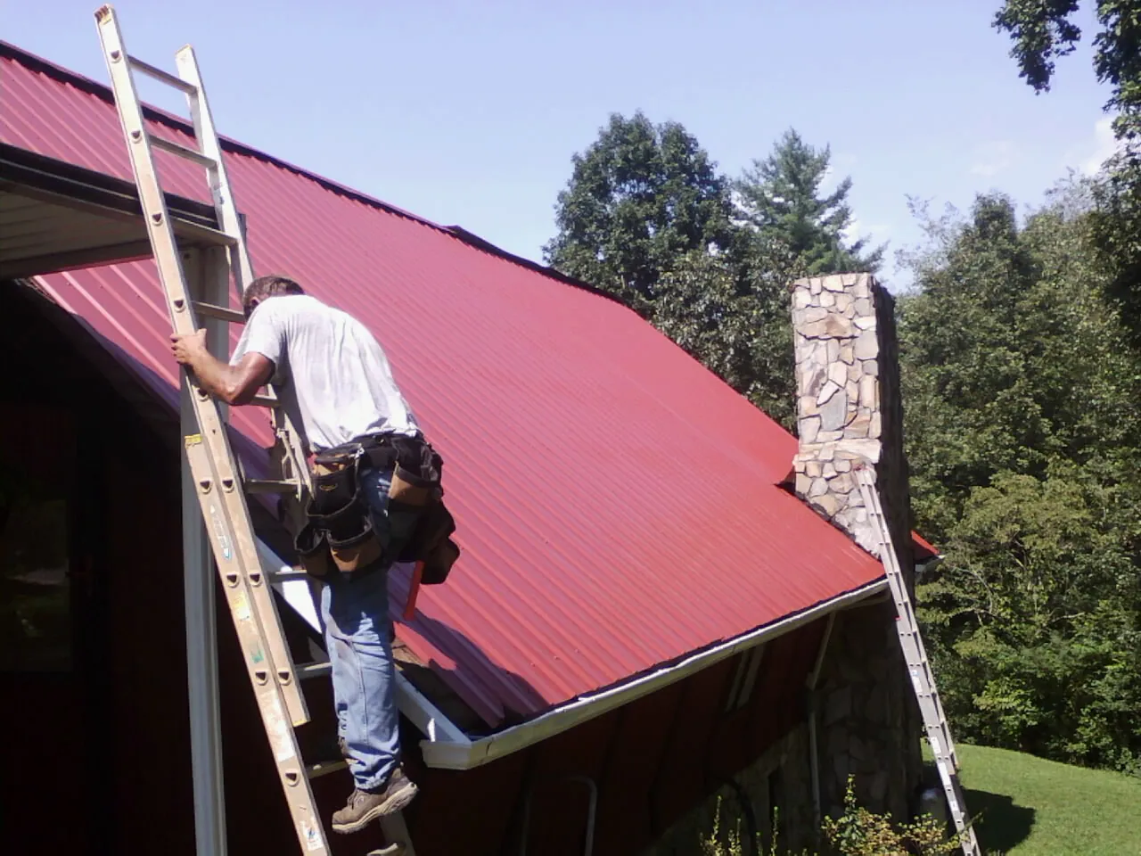 Roofer on a ladder beside a red metal roof and stone chimney — Soffit & Fascia Repair by Roofing Brothers in Jbsa Lackland